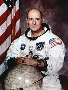 Astronaut Thomas P. Stafford, dressed in his space suit, with his arms resting on a globe, and an American flag is the background