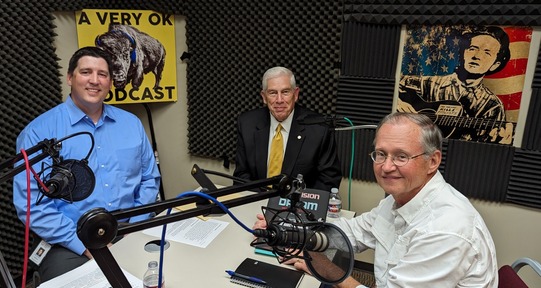 Trait Thompson, Wes Milburn, and Dr. Bob Blackburn seated in the recording studio. A Very OK Podcast and a Woody Guthrie poster can be seen.